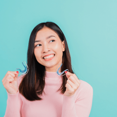 Teenage girl wearing long sleeved, pink shirt, looking up and smiling while holding orthodontic appliances, representing average orthodontic treatment time